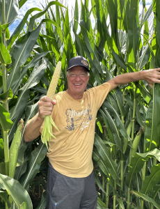 Autor sonriendo delante de sus plantas de maíz mientras sostiene un maíz en una mano.